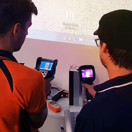 Two thermography students in a classroom aiming thermal cameras at a heated test rig during a practical exercise